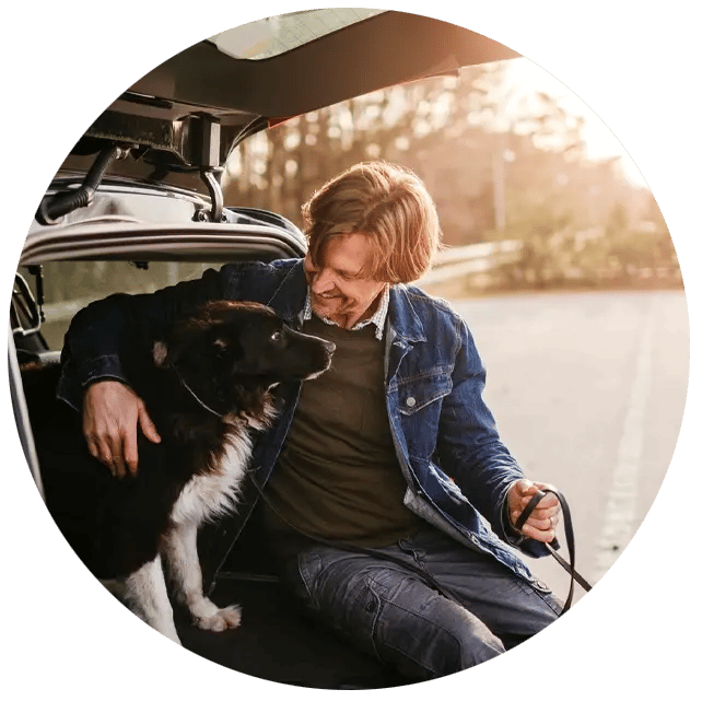Man with his arm around his dog, sitting in his open trunk. 